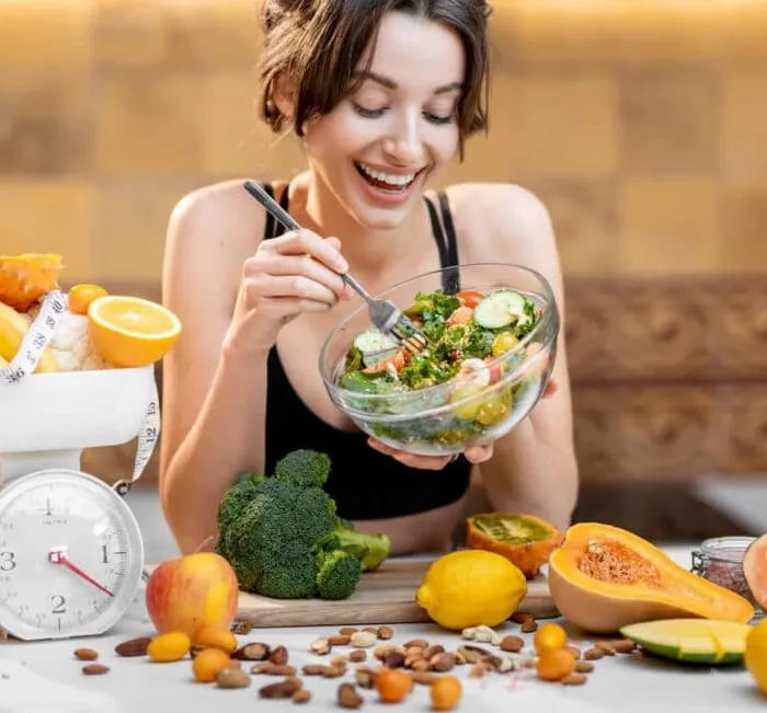 A smiling woman eats a bowl of salad at a table full of fresh vegetables and fruit